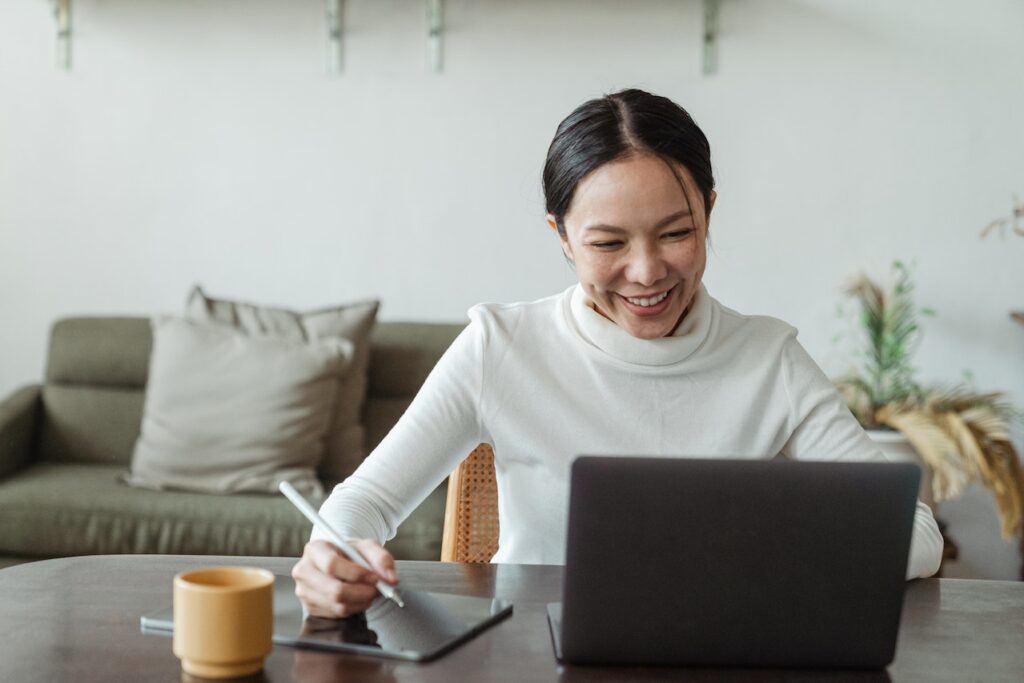 An Asian woman in a white turtleneck smiles as she works on a tablet and sits in a virtual meeting.