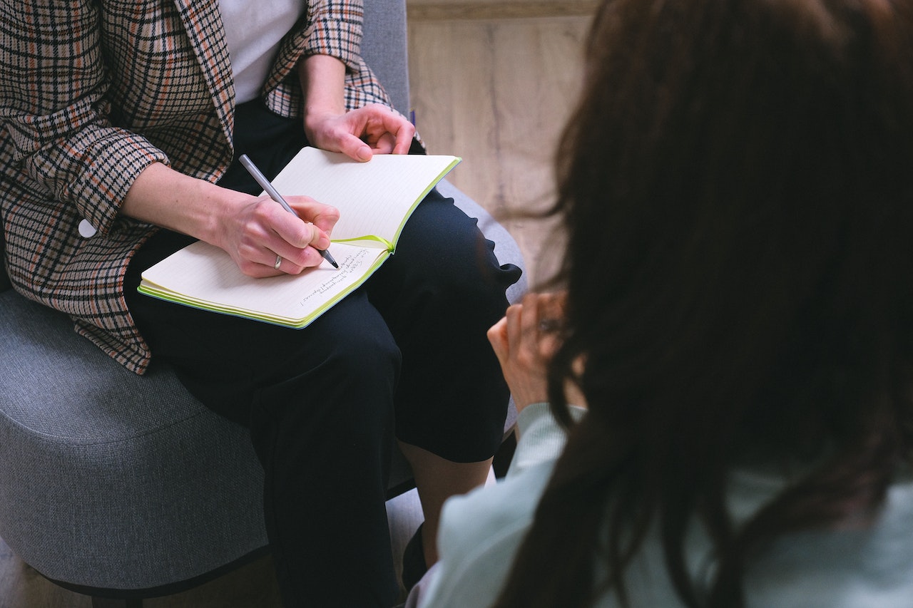 A seated woman in a plaid blazer takes notes as a client shares during a counseling session