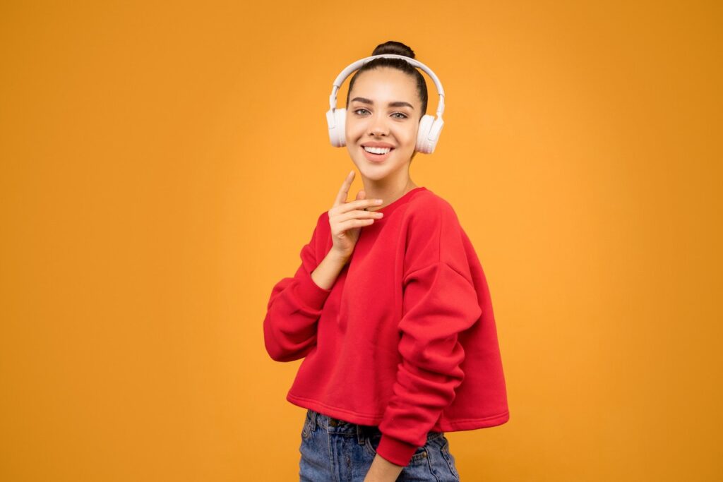 A lady with a bun wearing jeans, a red sweater, and white headphones poses against an orange background with her right hand touching her chin.