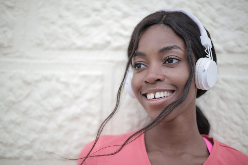 A black woman in a pink shirt smiling as she listens to something with her white headphones